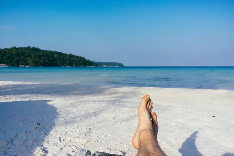Person lying back on the sand with their feet toward the sea on holiday, evoking the self-paced calm of Penthrox