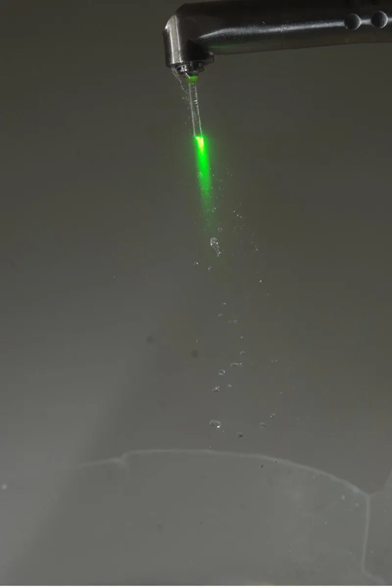 Real photograph of the Fotona dental laser handpiece emitting its green aiming beam through cascading water droplets — visually demonstrating how the Er:YAG wavelength is absorbed by water, the principle that makes it ideal for delicate soft-tissue procedures like frenectomy