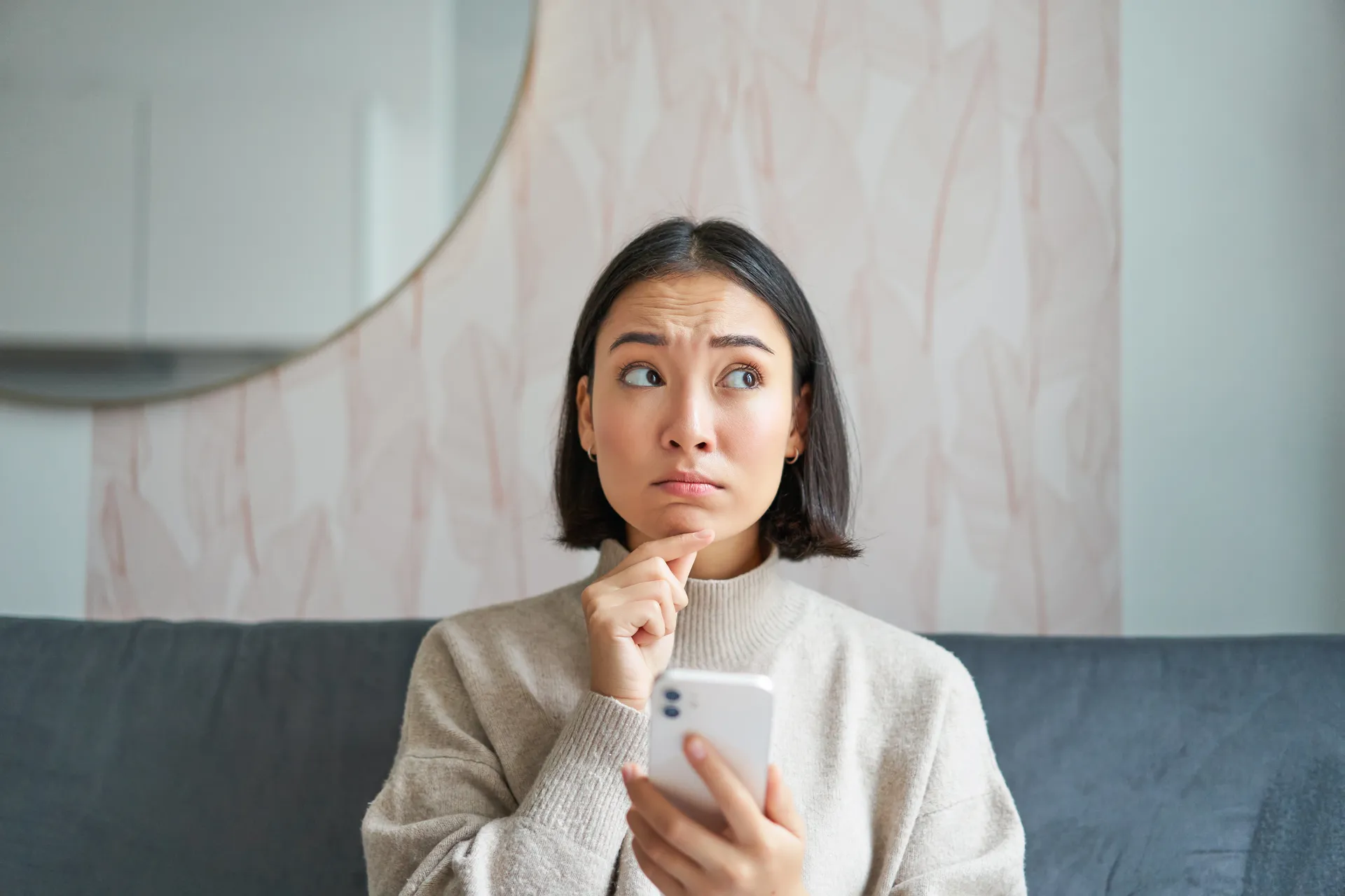 A woman sitting on her couch, holding her phone, hesitating before calling to ask about cost